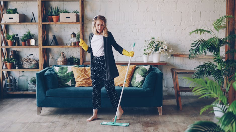 A woman with shoulder-length blonde hair, wearing a white T-shirt, a dark cardigan, and black polka-dot pants, is engaged in surface cleaning within a modern living room. She is barefoot, wearing yell