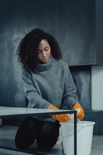 A woman with shoulder-length blonde hair, wearing a white T-shirt, a dark cardigan, and black polka-dot pants, is engaged in surface cleaning within a modern living room. She is barefoot, wearing yell